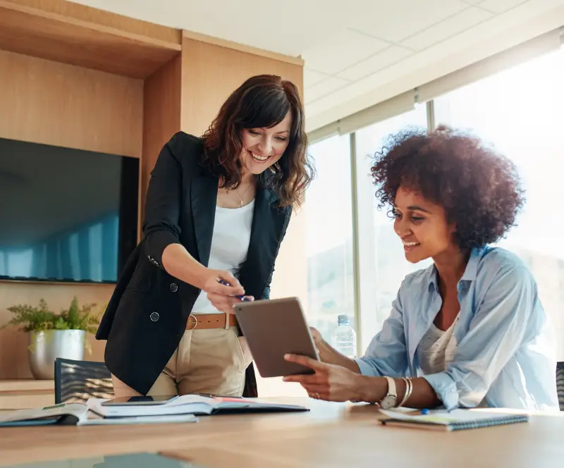 Image of a woman sitting at a desk and another woman pointing something out to her on her Ipad